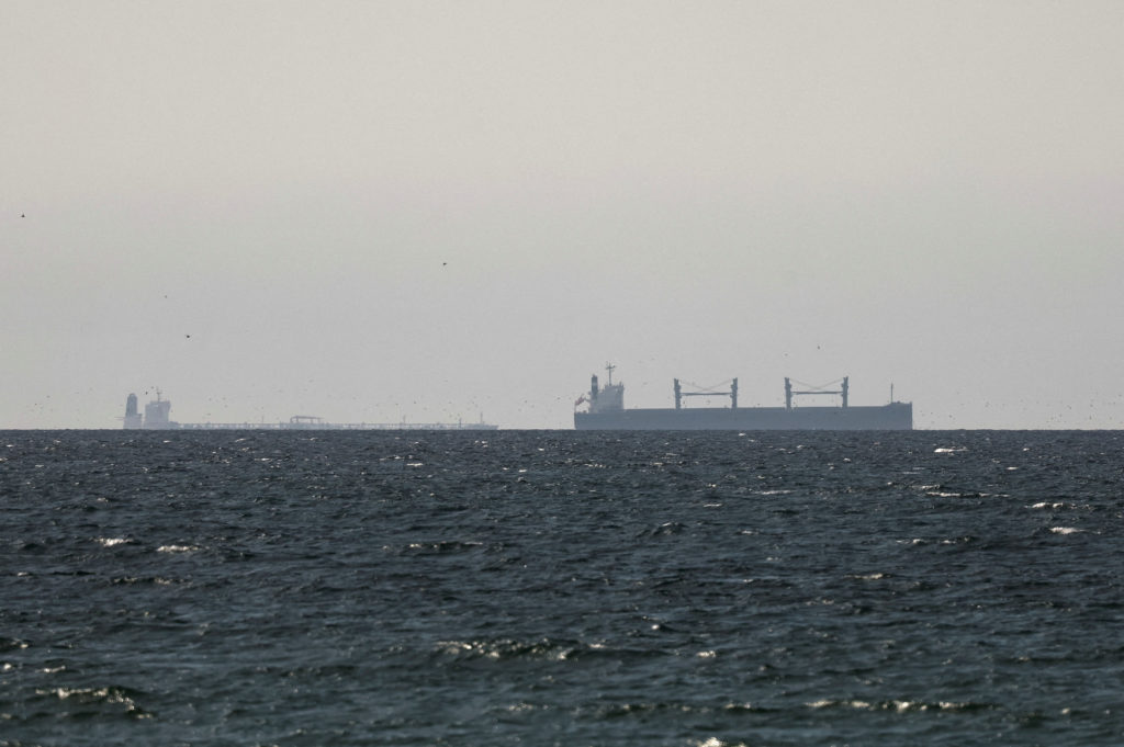 President Trump speaking at a podium with a map or graphic of the Strait of Hormuz and naval vessels in the background