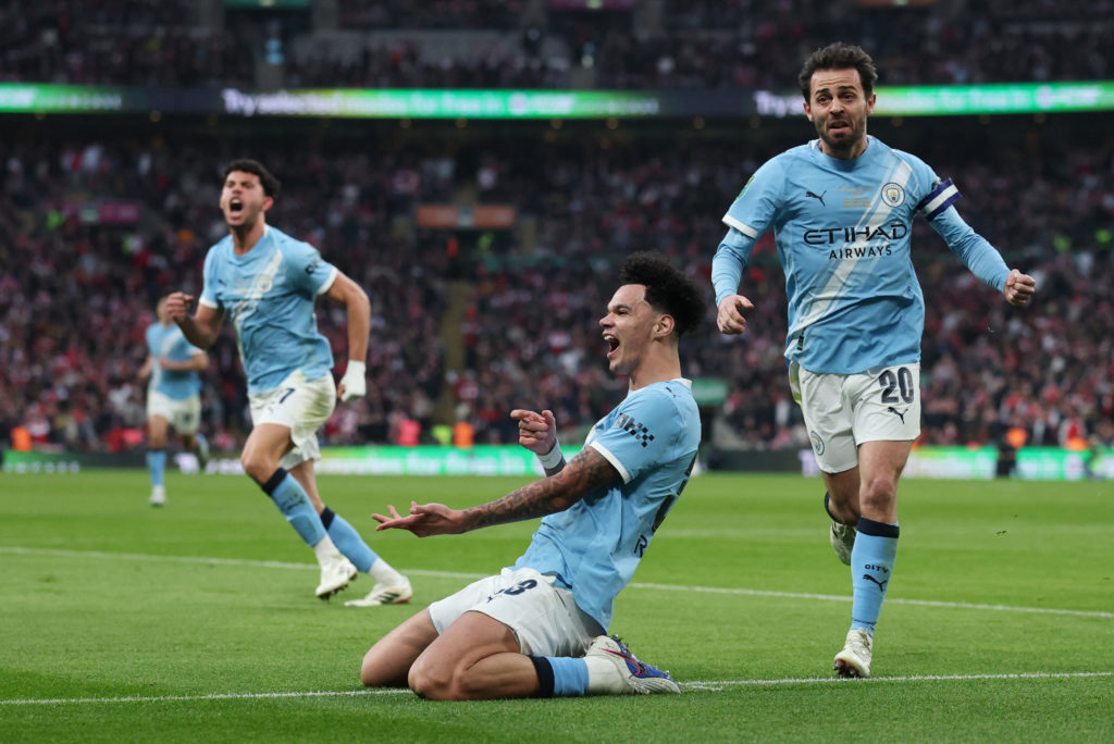 Manchester City players celebrating with the League Cup trophy on the pitch as confetti falls and fans watch from the stands
