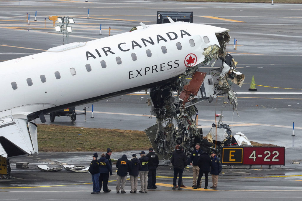 A damaged jet with its nose crushed beside a firefighting truck lying on its side on an airport ramp, scene at LaGuardia Airport