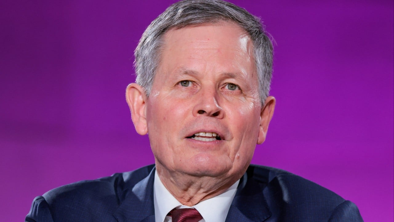 U.S. Senator Steve Daines in formal suit, with a blurred Hong Kong skyline in the background