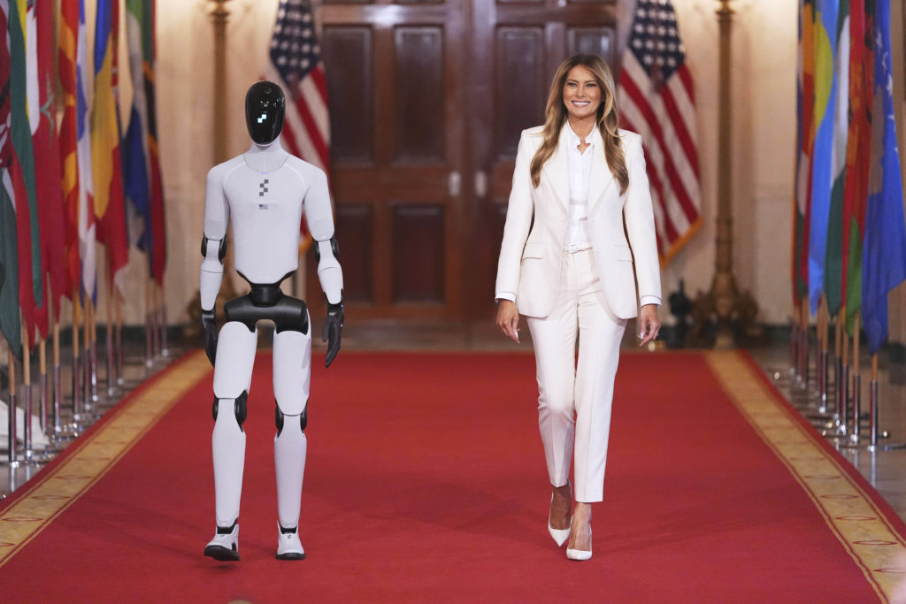 Melania Trump walking on a red carpet in a White House hallway alongside a humanoid robot near event attendees