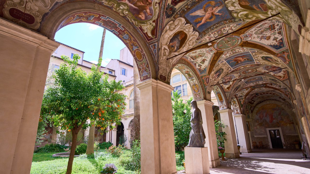 Interior view of a Roman cloister with a central pond, goldfish, turtles, frescoed walls and olive trees framed by two large palms