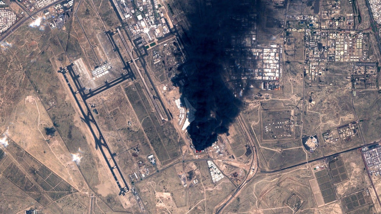 Smoke and flames rising from a fuel tank at an airport after a strike, with emergency responders at the scene
