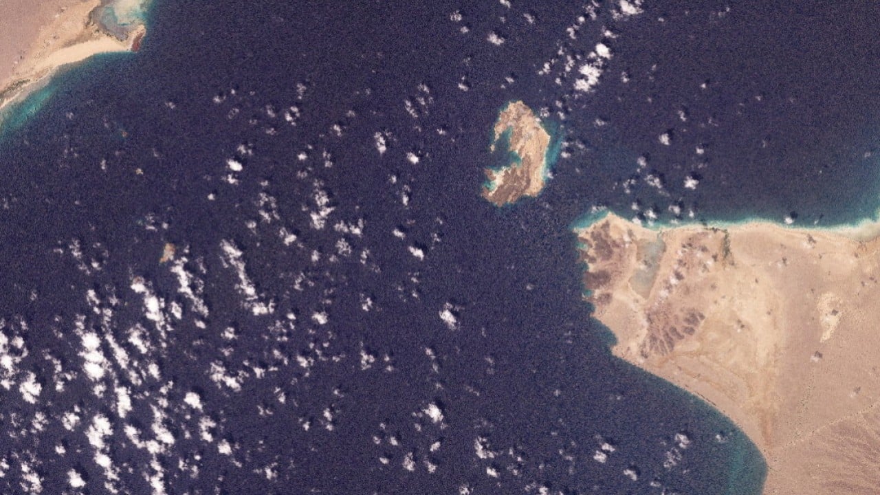 A cargo ship passing through a narrow Red Sea maritime chokepoint with military tension in the background