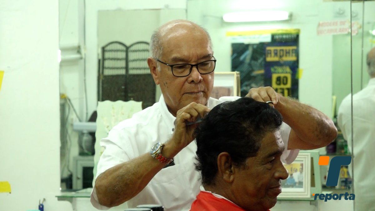 An elderly barber in David, Panama, standing beside a classic barber chair in a traditional barbershop