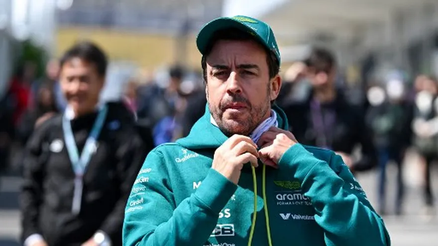 Fernando Alonso speaking to the media at a Formula 1 event with race cars and team garages in the background